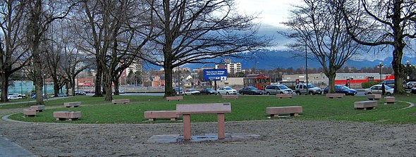 In a park, 14 coffin-like benches of pink stone are set in a circle. A higher slanted pink panel is visible in the foreground.