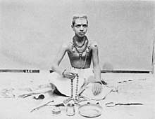 This image shows a young Hindu Priest sitting on floor with folded feet, "tilak" on his forehead, [[Rudraksh]] mala around his neck, [[Mala]] in his hand and other Pooja ritual necessities laying around him.