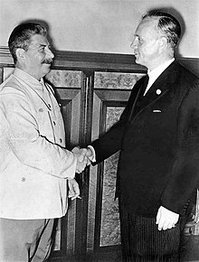 Two smiling men shake hands. Man on left wears a white suit and holds a cigarette. Man on right wears a black suit.