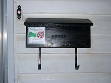 A black rectangular mailbox attached to the outside of a house. There is a doorbell above and to the left of the mailbox.