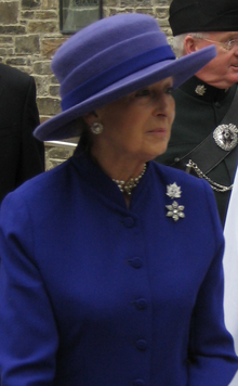 Photo en couleurs d'une femme vêtue de violet et portant un chapeau également violet avec un militaire en uniforme et un bâtiment de pierres en arrière-plan