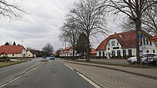 Straßenszene, d. h. auf der Straße, ein Fahrzeug rund 50 Meter voraus. Links Häuser, rechts Häuser und eine Bushaltestelle sowie Speisegaststätte „Selinus“