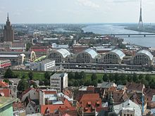 Farbfotografie in der Obersicht von vier großen Hallen im Rundbau mit vielen umliegenden Gebäuden. Eine lange Grünfläche erstreckt sich im vorderen Bereich. Im linken Hintergrund ist ein brauner Wolkenkratzer und in der hinteren Mitte geht eine Flussabzweigung vom rechten großen Fluss. Rechts verbindet eine Brücke das hintere Ufer, auf dem ein riesiger, moderner Turm steht, mit dem vorderen Ufer.