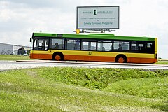 Autobus komunikacji gminnej. Pomalowany w poziome pasy o kolorach: zielonym, żółtym i pomarańczowym. Na dole zdjęcia trawa. W tle bilbord.