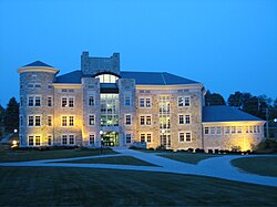 A three-story stone building with a round tower structure on the left and a large window structure in the middle, with brick walkways leading to the entrance from several directions