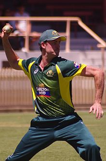 A cricketer dressed in a green uniform with a cap about to throw a cricket ball with his right hand towards the right of the photograph