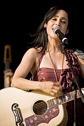 A woman in a purple dress singing to a microphone and playing the guitar.