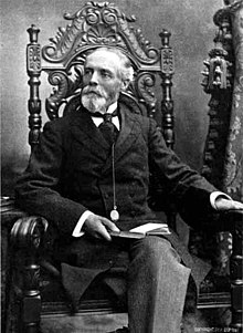 Seated portrait of 60-ish man with beard, mustache, and receding hairline, in circa-1880 formal dress with high collar, with a book in his lap and a watch at the end of a chain suspended from his coat.