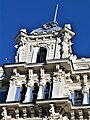 Frontale Farbfotografie in der Untersicht von einer oberen Fassade mit einer Dachhaube, deren Spitze ein langer Stab ist. An den Ecken des oberen Fenstervorbaus befinden sich zwei Figuren und zwischen den unteren Fenstern stehen Säulen. Über den Fenstern sind Ornamente.