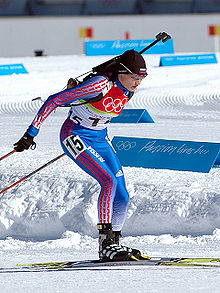 Photographie d'une biathlète en course, vêtue d'une combinaison bleue et rouge, portant des lunettes et les bâtons en arrière.