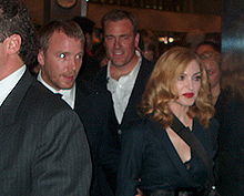 Image of a group of people. Amongst them a woman is prominent. She is wearing a black dress and has deep, red lips. Her hair is light brown in lcolor and falls is waves around her face. She is looking to the left. To her right, only the face of a middle-aged, clean shaven man is visble, being obstructed by the left profile of the man in front whose shoulder is the only part visible.