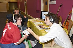 People gathered around a table wearing headphones. The journalist holds the microphone for a physically disabled person to speak