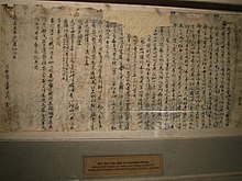 Morceau de papier abîmé sur lequel on lit des écritures en caractères chinois écrits à l'encre sombre. Le papier est encadré et exposé dans un musée.