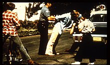 Photo of a person being arrested in the street by a policeman. The policeman is fixing the person's arms behind his back and pushing him towards a police car. One person kneeling at the side of the car is taking pictures. Two children in the foreground are watching the scene.