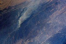 Viewed at a steep angle from above, a plume of gray smoke rises and drifts across parched brown mountains