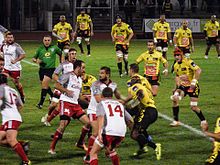 Quelques joueurs de rugby de deux équipes se regroupent, tandis que les autres joueurs occupent la surface du terrain.