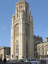 A tall stone nineteenth century with shields on the visible sides and a pepperpot upper storey. In front, traffic and pedestrians on a busy street.