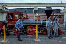 A red steam locomotive with a 4-4-0 wheel arrangement (four leading wheels, four driving wheels, and no trailing wheels) passing over a bridge