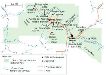 A large green area representing Chaco Culture National Historical Park's boundaries sits in the middle of a white field. The green area is roughly rectangular with one smaller square-like and one triangular appendage abutting it at bottom-left and bottom-right, respectively. Fifteen small red circles represent the location of important Chacoan sites; they are focused on a line running from top-left (northwest) to bottom-right (southeast). A dashed blue line depicting the Chaco Wash runs roughly along the same line; a network of dashed and solid orange lines represent trails and metalled roads, respectively, also focus on the same axis, connecting the red dots. Two gold squares define high points: "Fajada Butte (2019 m.)" and "West Mesa (2035 m.)".