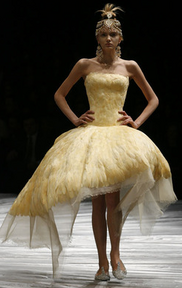 Woman wearing gold jewelled headdress and butter yellow sleeveless dress with straight neckline and knee-length asymmetrical puffy skirt. The yellow part of the dress is made of feathers, with white or sheer tulle underneath. She has flat shoes.