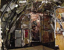 Photo couleur prise à l'intérieur d'une reconstitution de l'avion dans un hangar. Les débris métalliques sont placés sur des grilles correspondant à leur emplacement sur l'avion.