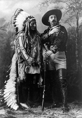 Black-and-white studio photograph of Sitting Bull and Buffalo Bill, standing in front of a forest backdrop. Buffalo Bill towards an imaginary horizon.