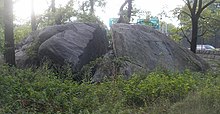 A photograph of a very large rock, about the size of a small truck, that has a large fissure in the middle. The rock is surrounded by trees and other vegetation.