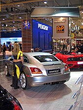 Une voiture 2 portes grise vue de l'arrière gauche exposée dans un salon aux côtés d'autres automobiles. Une femme s'appuie sur la portière conducteur de la voiture.