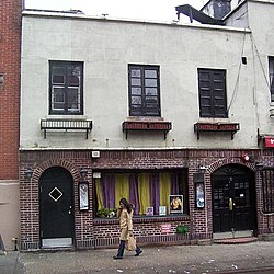 The facade of 53 Christopher Street as seen from across the street. The facade is two stories high. At the ground or first story, the facade is made of brick and consists of a wide window flanked by two archways. At the second story, there is a stucco facade with three windows, each with a flower-pot holder below.