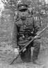 A man in a military uniform is kneeling on one knee. He is armed with a rifle and a hand grenade. A light forest is behind him.