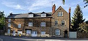 A row of English stone houses with white windows and doors beside a road. There is a stone wall and wooden gate on one side, and several different types of trees around them.