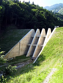 Na fotografii je hráz umístěná v údolí. Hráz je částečně otevřená, mohutná mříž z betonu brání v průchodu velkým objektům.