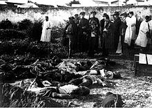 Photograph of several dead bodies lying on the floor. Above them stand members of the police and reporters.