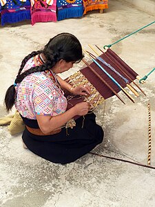 A loom made of sticks and string. The top endbar is tied to a fixed object using green rope; the lower end bar is attached to a leather strap around the weaver's back. Between, two heddle rods and several shedding rods. The sticks to one side are probably sword beaters. No shuttles or bobbins are being used.