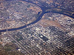 High angled aerial shot of a developed city; a suburban grid dominates the lower half of the image. A river bisects the city from the left before forking; the first fork continues up and to the right edge of the image; the second curves up and around to finish on the left, enclosing industrial units and other domestic properties.