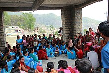 Sous un abri en pierre et en bois, assemblée de jeunes filles népalaises assises par terre en tenue bleue, assistant à un cours de sensibilisation.