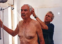 A man steadies himself by holding a grab bar in the shower, while another man shampoos his hair