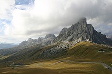 Gunung yang ditutupi awan menjadi latar belakang lembah hijau di bawahnya, dengan jalan serpentin melaluinya.