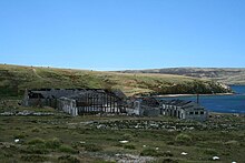 A cluster of ruined industrial buildings sits on the shore of a green, treeless landscape.