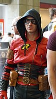 Photo d'un homme portant un costume rouge, noir et doré avec sa capuche sur la tête.