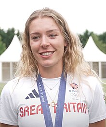 Eve Stewart trägt ein weißes T-Shirt mit der Aufschrift Great Britain. Um den Hals hängt ein blaues Band, an dem ihre olympische Medaille befestigt ist, die allerdings nicht im Bild zu sehen ist.