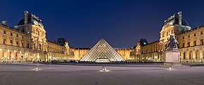 Frontale Nachtaufnahme von einem breiten Platz mit einem beleuchteten Barockschloss aus drei Flügeln. In der Mitte des Platzes steht eine beleuchtete Glaspyramide und an beiden Schlossseiten sind zwei kleine Pyramiden. Links und rechts sind vier Anbauten mit einem weiteren Geschoss und verzierten Dachkuppeln sowie Säulenfassaden. Im Erdgeschoss sind überall Arkaden und über vielen Fenstern sind Giebelfelder. Rechts vorne ist auf einem Podest eine Reiterfigur.