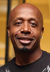 A image of a black man with an earring in his left ear. He is smiling and wearing a brown shirt.