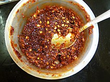 A red spread containing seeds in a white bowl, viewed top-down, with a metal spoon inside.