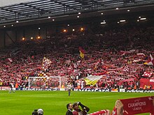 A single-tiered stand that contains thousands of people. Several flags are being waved. In front of the stand is a grass pitch with a goal.