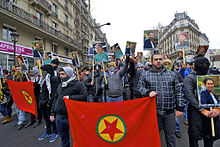 A group of people marching down an urban street behind a red flag with a star in the middle. They are holding up placards with pictures of three different women on them