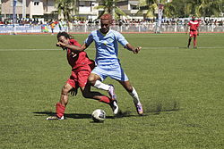 Photographie d'une scène d'un match de football : un joueur en bleu bouscule un autre joueur rouge pour le ballon.