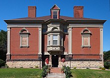 A light-red-brick building. A bay window sits above the entrance, while two stone signs to the left and the right, respectively, read "Nelson" and "A.D. 1837". The building has two chimneys and a window at the top middle.