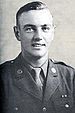 Head and shoulders of a young white man with neatly combed hair wearing a military jacket with a round pin on each lapel over a shirt and tie.