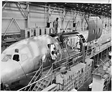 photo noir et blanc, chaîne d'assemblage, un grand panneau de fuselage est amené par une grue.